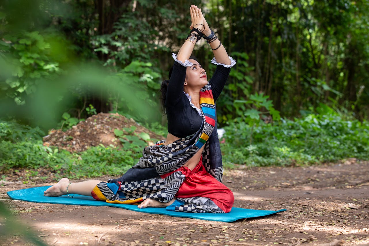 Woman practicing yoga pose outdoors in vibrant traditional attire, surrounded by lush greenery.